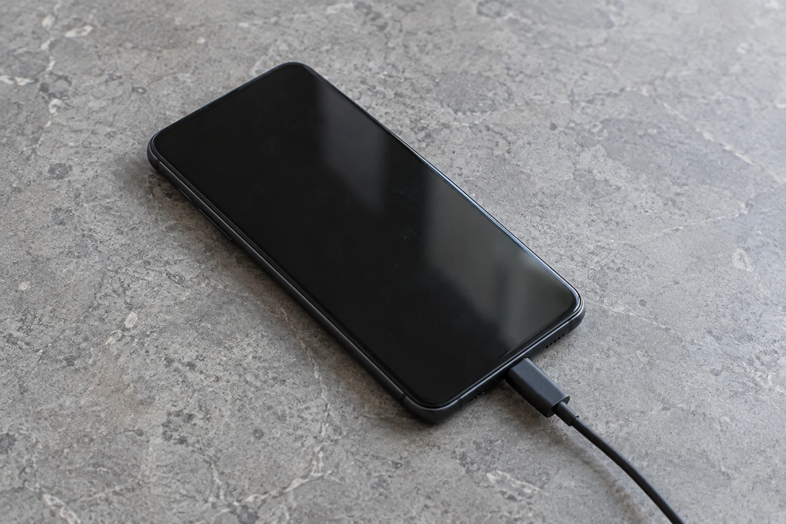A smartphone connected to a charging cable with its screen off on an open grey counter